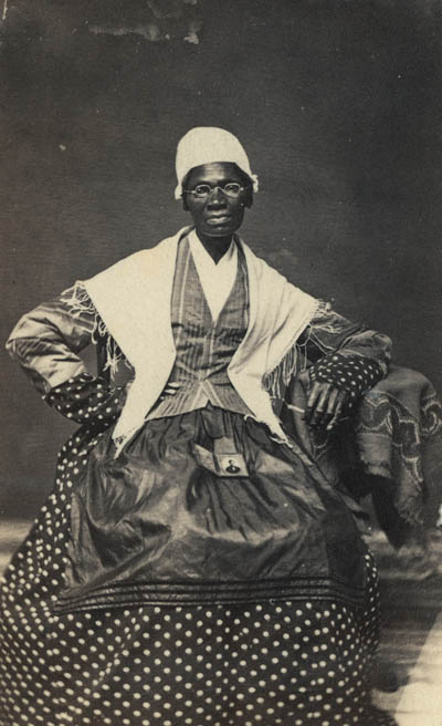 <p>Black and white photograph of a middle aged Black woman wearing glasses, a white cap and shawl, a polka-dotted dress, and a jacket and apron. She sits facing the camera with one hand on her hip and her other elbow resting on a small table. In her lap is a small framed photograph of her grandson.</p>